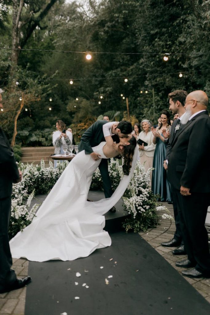 A couples romantic kiss at an outdoor wedding ceremony surrounded by guests and flowers.
